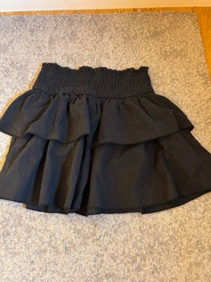Svart volangkjol med smock - Supersöt svart kortkjol med bred smock i midjan och dubbla volanger. Kjolen har en luftig känsla och passar perfekt till en trendig vardagslook. Ingen synlig logga eller märke. Materialet känns som bomull eller polyester.