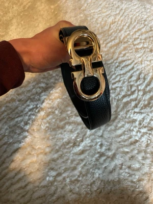 Svart skärp från Salvatore Ferragamo - Snyggt svart skärp från Salvatore Ferragamo med exklusivt guldfärgat spänne i metall. Bältet är gjort i präglat skinn och har den ikoniska dubbla Gancini-detaljen framtill. Perfekt accessoar för att lyfta din look.