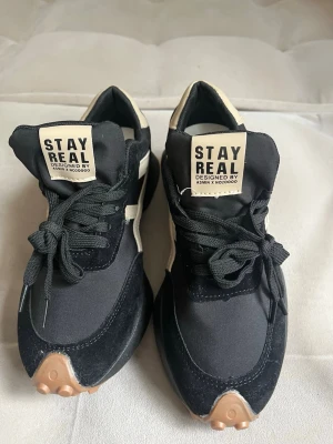 Svarta Stay Real sneakers med beige detaljer storlek 38/39 - Snygga svarta sneakers från Stay Real med beige detaljer och grov sula. Skorna har svart ovandel i textil och mocka, beige logga och häl samt bruna detaljer på sulan. Perfekta för en trendig streetstyle-look. Helt nya storlek 39 men passar 38 lite små för mig som har 38.5 -39