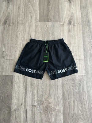 Svarta shorts från BOSS med logga - Svarta shorts från BOSS med elastisk midja och vit BOSS-logga tryckt längs nederkanten. Perfekta för en avslappnad och sportig look. Tillverkade i ett lätt och bekvämt material.
