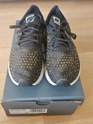 Nike svarta och guldiga sneakers - Snygga Nike sneakers i svart med guldiga detaljer och vit sula. Ovandelen är i meshmaterial som andas och har ett guldigt Nike-märke på sidan. Skorna har klassisk låg profil och snörning framtill. Perfekta för dig som vill sticka ut med stilrena detaljer. Använda ett fåtal gånger. Nypris 1300 kr