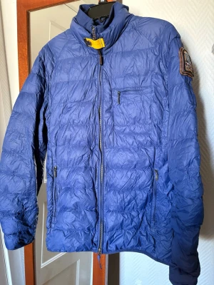 Blå dunväst från Parajumpers - Snygg blå dunväst från Parajumpers med hög krage, dragkedja framtill och flera fickor med dragkedja. Märkets ikoniska patch på ärmen och gul detalj vid halsen. Perfekt för lager-på-lager och håller dig varm med lättviktig dunfyllning. Små skador på armarna men de är lagade.storlek xl men skulle säga large