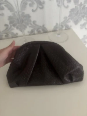 Mörkbrun flätad clutchväska - Snygg mörkbrun clutchväska i flätad look med mjuk form. Väskan har guldfärgade detaljer på insidan och rymligt fack. Perfekt för dig som gillar stilrena accessoarer med unik textur. Passar till många olika outfits och är lätt att bära med sig.