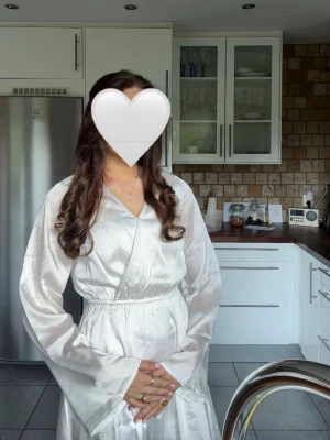 Vit skolavslutnings klänning satin - Snygg vit omlottklänning i satin med långa ärmar och markerad midja. Klänningen har en elegant v-ringning och ett mjukt, glansigt material som ger en lyxig känsla. Perfekt för dig som gillar stilrena och trendiga plagg.