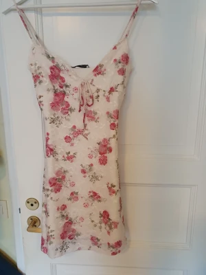 Blommig spetsklänning från Bershka - Supersöt kort klänning från Bershka i ljus spets med rosa och röda blommor. Klänningen har smala axelband, snörning framtill och en figurnära passform. Perfekt för sommarens alla tillfällen!