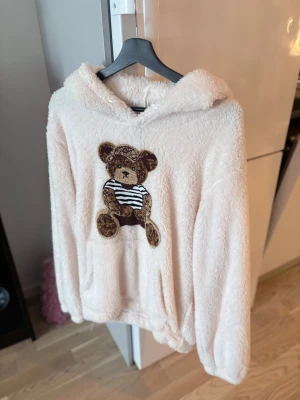 Ljus fluffig hoodie med nallemotiv - Supermjuk och fluffig hoodie i ljusbeige med stor broderad nalle på bröstet. Nallen har randig tröja och bruna detaljer. Hoodien har luva och magficka, perfekt för kalla dagar. Materialet är teddytyg som känns riktigt gosigt.