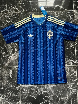 Blå Sverige fotbollströja Adidas - Säljer en blå fotbollströja från Adidas med Sveriges landslagsemblem på bröstet. Tröjan har ett unikt mönster i olika blå nyanser, ljusgula detaljer på axlarna och V-ringad krage. Perfekt för match eller träning!