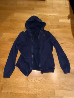 Två Mörkblåa hoodies från Lyle & Scott - Två Mörkblåa tjocktröjor en med dragkedja och en utan från Lyle & Scott. Ena är för 14-15 år och den andra 15-16år. Dem är ganska små i storlek. Klassisk modell med huva, två fickor framtill och gul logga på bröstet. Perfekt för en avslappnad och stilren look. Mjuk bomullskänsla och ribbade muddar vid ärmslut och nederkant.