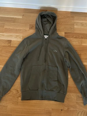 Olivgrön hoodie från Lacoste - Snygg olivgrön hoodie från Lacoste med dragkedja framtill, två stora fickor och klassisk krokodillogga på bröstet. Huva med dragsko och ribbade muddar vid ärmslut och nederkant. Perfekt för en chill och stilren look.