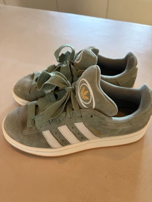 Gröna Adidas sneakers i mocka - Snygga gröna Adidas sneakers i mocka med klassiska vita ränder på sidorna och gummisula. Skorna har breda gröna skosnören och Adidas logga i vitt och gult på plösen. Perfekta för dig som  gillar streetwear. Är i superbra skick! 