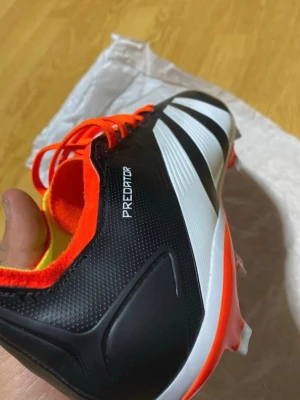 Adidas Predator fotbollsskor svart/vit - Snygga Adidas Predator fotbollsskor i svart och vitt med klassiska tre ränder och orange detaljer. Skorna har syntetmaterial, snörning och låg profil för bra bollkontroll. Perfekta för dig som vill sticka ut på planen.