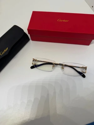 Glasögon från CARTlER - Säljer ett par CARTlER CT00920 (Piccadilly) från kollektionen Signature C. Dessa bågar är symbolen för sofistikerad minimalism och hantverk av högsta kvalitet. Detaljer: * Modell: Randlös (rimless) design som ger ett lätt och luftigt intryck. * Material: Detaljer i högblank guldfinish med kontrasterande svarta skalmar. * Skick: Oanvända * Tillbehör: Säljes komplett med box samt förvaringslåda Nypris: 10 000-12 000 kr Finns för avhämtning i Stockholm eller skickas spårbart och försäkrat via plick