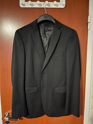 Dressmann kostymkavaj - Stilren svart kavaj från Dressmann i slim fit-modell. Kavajen har klassisk passform med två framfickor med lock, bröstficka och snyggt foder med röda detaljer på insidan. Perfekt för dig som vill ha en modern och enkel look.