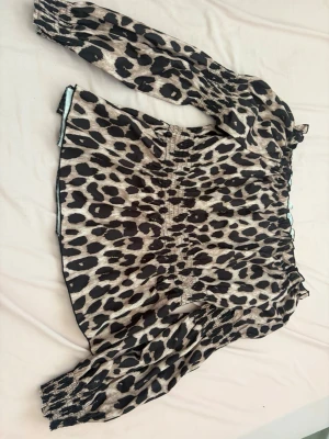 Leopardmönstrad blus med puffärm - Snygg blus med leopardmönster i beige och svart. Modellen har långa puffärmar med resår vid ärmslut och smockad midja för en figurnära passform. Tunt, lätt material som ger en luftig känsla. Perfekt för dig som vill sticka ut med ett trendigt djurmönster.