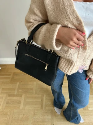 Svart handväska med gulddetaljer🌸💖💖 - Stilren svart handväska i slätt konstläder med guldfärgade dragkedjor och detaljer. Väskan har ett ytterfack med dragkedja och två korta handtag, perfekt för att bära i handen eller på armen. Enkel och klassisk design som passar till många outfits.