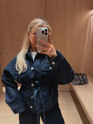 Mörkblå jeansjacka med bälte Bershka - Mörkblå jeansjacka från Bershka med knappar, två bröstfickor och tillhörande bälte i midjan. Jackan har en klassisk krage och raka ärmar