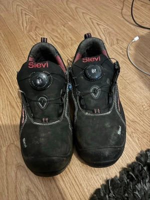 Arbetsskor Sievi med BOA-snörning - Svarta sneakers från Sievi med BOA-snörning för enkel justering. Skorna har rosa detaljer och vit kontrastsöm, samt en vadderad plös och insida i rosa mesh. Ovandelen är i slitstarkt material och sulan är robust för extra komfort. Har även stålhätta. 