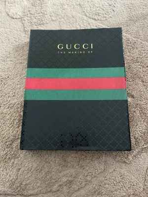 Gucci: The Making Of bok - Stilren och lyxig bok med titeln 'Gucci: The Making Of'. Omslaget är svart med klassiska gröna och röda ränder samt guldfärgad text. Perfekt för modeintresserade och som coffee table-bok. Hård pärm och exklusiv design. Har en skavank mellan sida 1-5 se bild 5 annars hel och ren på omslaget och utsidan 