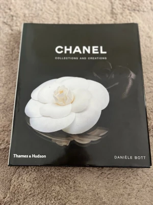 Chanel Collections and Creations bok - Snygg bok med svart och vit hård pärm, prydd med en stor vit blomma på omslaget. Boken är skriven av Danièle Bott och handlar om Chanels ikoniska mode och design. Perfekt för dig som gillar mode, design och lyxiga varumärken. Snygg som prydnad eller coffe tabel book 