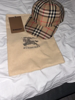 Burberry keps med klassiskt rutmönster - Snygg keps från Burberry i deras ikoniska beigea rutmönster med inslag av svart, vitt och rött. Kepsen har böjd skärm och justerbar rem bak. Perfekt accessoar för att lyfta din streetwear-look.