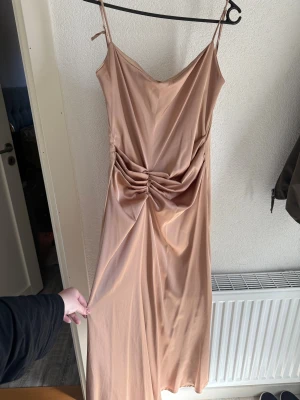 Champagnefärgad långklänning Label Nue - Snygg långklänning från Label Nue i champagnefärgad satin med smala axelband och v-ringad front. Klänningen har en elegant rynkning framtill som ger en cool och modern look. Perfekt för dig som vill sticka ut med stil och komfort.