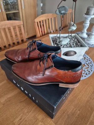 Bruna loafers från Bugatti i skinn - Snygga bruna loafers från Bugatti i äkta skinn med mörkblåa detaljer och snörning. Skorna har en klassisk design med låg klack och bekväm innersula. Perfekta för dig som vill ha stilrena och bekväma skor till vardagslooken.