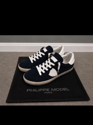Philippe Model mörkblå vittsnöre sneakers - Snygga sneakers från Philippe Model i mörkblå mocka med vita detaljer och vit sula. Klassisk låg modell med vita skosnören och logotyp på sidan. Perfekta för dig som gillar stilrena och trendiga skor med premiumkänsla. Kvitto finns. Utanbox.