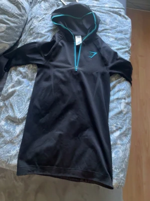 Onyx hoddie helt ny - Svart hoodieklänning från Gymshark med turkosa detaljer och halv dragkedja framtill. Tillverkad i en mix av polyester, nylon och elastan. Perfekt för träning eller chill, med sportig look och luva. Helt ny 