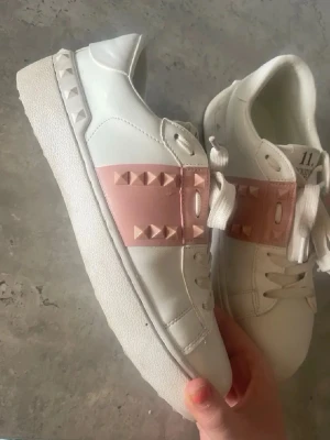 valentino skor - Snygga vita sneakers från Valentino med bred rosa rem över snörningen dekorerad med matchande pyramidformade nitar. Skorna har en chunky vit sula och klassisk låg profil. Tillverkade i slätt läder med vita skosnören och coola detaljer på hälen. säljer för att dom är för små💕