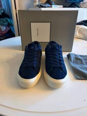 Tom Ford skor - Snygga sneakers från Tom Ford i mörkblå mocka med vit sula och klassisk låg profil. Skorna har diskret branding på hälen och matchande blåa skosnören. Perfekt för dig som vill ha en stilren och exklusiv look.