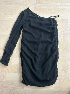 Svart oneshoulder klänning - Svart kortklänning med oneshoulder-design och lång ärm. Klänningen har rynkade detaljer längs sidan och är tillverkad i ett lätt, något transparent material med diskret struktur. Perfekt för dig som vill sticka ut med en unik siluett. Stl 158