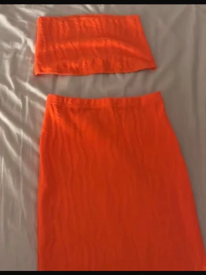  tvådelat set - Säljer ett orange tvådelat set med en bandeau-topp och en matchande kjol. Båda delarna har en snygg strukturerad yta med vågiga mönster. Setet är figurnära och stretchigt, perfekt för dig som gillar färgstarka plagg. Perfekt till sommaren!