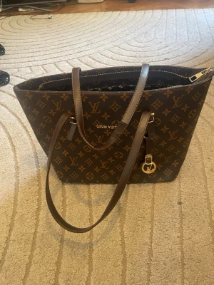 Superfin väska❤️ - Snygg brun handväska  med klassiskt monogrammönster i guld och bruna nyanser. Väskan har två långa axelband, guldfärgade detaljer och en praktisk dragkedja. Perfekt för dig som vill ha en lyxig och trendig accessoar. Lite slitna handtag