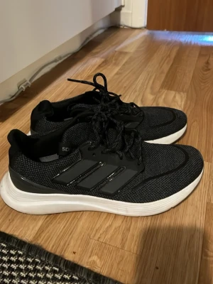 Svarta Adidas sneakers med vita sulor - Snygga svarta Adidas sneakers med klassiska tre ränder på sidan och vit, tjock sula. Ovandelen är i meshmaterial som andas och skorna har svarta snören. Perfekta för en sportig och stilren look.
