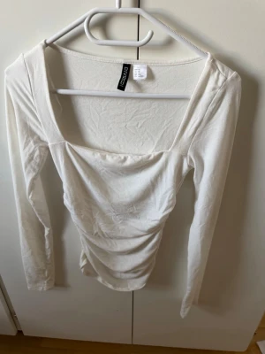 Vit långärmad topp från Divided - Snygg vit långärmad topp från Divided med fyrkantig ringning och lätt rynkade detaljer framtill. Tillverkad i mjuk bomull som känns skön mot huden. Perfekt att styla med jeans eller kjol för en trendig look.
