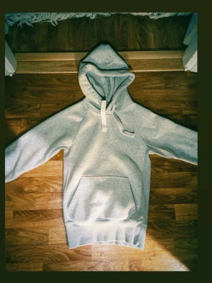 Ljusgrå hoodie med magficka -  Använt bara 2 gånger. Snygg ljusgrå hoodie med stor huva och breda vita dragsnören. Hoodien har en klassisk magficka framtill och ribbade muddar vid ärmslut och nederkant. Perfekt för en avslappnad och stilren look.