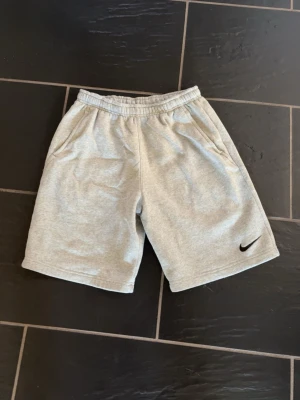 Grå sweatshorts från Nike - Grå mjuka sweatshorts från Nike med elastisk midja och två sidofickor. Klassisk svart Nike-logga broderad på vänster ben. Perfekta för avslappnade dagar eller träning. Tillverkade i bomullsmix för skön komfort.