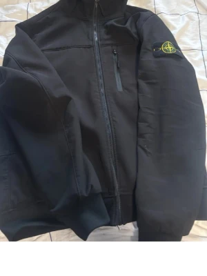 Svart jacka från Stone Island - Säljer en svart jacka från Stone Island med dragkedja framtill och bröstficka med dragkedja. Klassisk patch med logga på vänster ärm. Jackan har en stilren design och ribbade muddar vid ärmslut och nederkant. Perfekt för dig som gillar streetwear.
