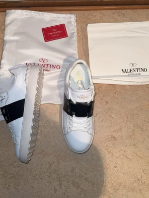 Valentino Garavani vita sneakers - Snygga vita sneakers från Valentino Garavani med bred svart rem över vristen. Skorna är i läder med klassisk snörning och har en chunky sula med mönstrad undersida. Perfekta för dig som vill ha en stilren och lyxig look.