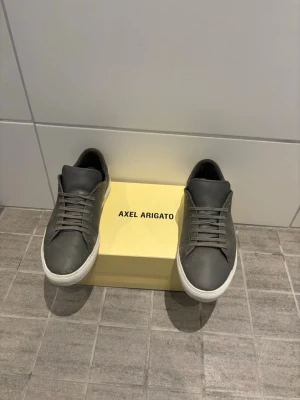 Gråa Axel Arigato sneakers i läder - Snygga grå sneakers från Axel Arigato i stilren design. Skorna är tillverkade i slätt läder med matchande grå skosnören och vit sula. Perfekta för dig som gillar minimalistisk och clean stil. Klassisk låg modell som passar till det mesta.