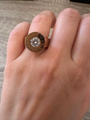 Rosefärgad ring med stor sten från LenMer - Cool rosefärgad ring med en stor, glittrande sten i mitten. Ringen har en bred design och text ingraverad runt stenen. Perfekt statement piece för dig som gillar att sticka ut. Ringen är i storlek 6 och helt oanvänd! OBS: FRAKTAR INOM 24H!🩷