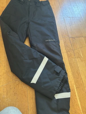 Svarta täckbyxor med reflexdetaljer - Svarta täckbyxor från Cross Sportswear med raka ben och breda reflexband vid bensluten. Byxorna har justerbar midja och är tillverkade i ett slitstarkt, vattenavvisande material. Perfekta för kalla dagar och utomhusaktiviteter.