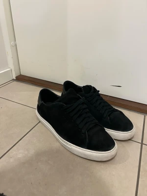 Axel Arigato  - Stilrena svarta sneakers med vit sula och svarta skosnören. Ovandelen är i mockaliknande material och hälen har en detalj i blankare svart. Perfekta för en clean och enkel look.