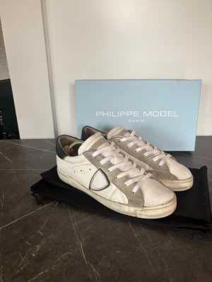 Philippe Model  - Snygga vita sneakers från Philippe Model med gråa och beige detaljer. Skorna är i stl 40. Men är stora i stl vilket Philippe Modells är. Skorna har använts flitigt och har några defekter. På bild 6 syns en tydlig defekt. Bortsett från det så är det en perfekt sko för dig som vill gå runt med stil! ”Skorna köptes från Merchsweden.se” dvs 100% äkta. Dustbag och låda ingår! Är du intresserad i fler bilder så hör av dig i DM!  