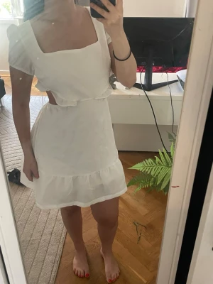 Vit kort klänning med cut-outs - Söt vit kort klänning med fyrkantig ringning, puffärmar och volangkant nertill. Klänningen har trendiga cut-outs i midjan och är tillverkad i ett lätt tyg, perfekt för varma dagar.
