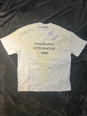 Vit Acne Studios t-shirt med tryck - Populär M vit t-shirt från Acne Studios  med svart texttryck 'STOCKHOLM 1996' på bröstet. Klassisk rak passform och rund halsringning. Tillverkad i mjuk bomull, perfekt för en clean och stilren look. OBS: osäker på äktheten då jag köpte den av en kompis, därför det låga priset (449kr ist för ca 2000-3000kr) Tveka inte att skriva om du har några funderingar!