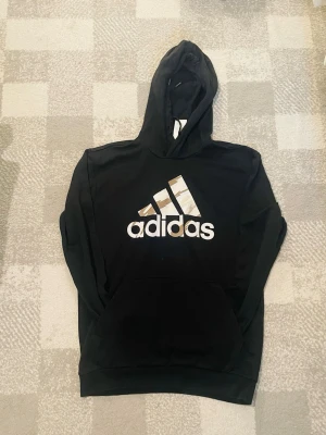 Svart hoodie från Adidas med logga - Snygg svart hoodie från Adidas med stor logga i vitt och guld på bröstet. Klassisk känguruficka framtill och huva. Perfekt för dig som gillar streetwear och sportig stil. Mjuk och bekväm bomullsblandning.