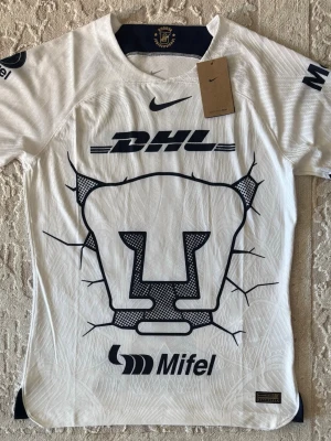 Vit Pumas UNAM matchtröja Nike - Säljer en vit matchtröja från Pumas UNAM med stor blå logga på bröstet och flera sponsortryck. Tröjan har korta ärmar, rund hals och är tillverkad i ett lätt, tekniskt material som andas. Perfekt för fotboll eller träning. 