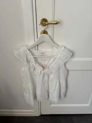 Ginatricot - Supersöt vit blus från Gina Tricot med volangdetaljer längs ringningen. Ärmlös modell i mjukt bomullstyg, perfekt för varma dagar. Passar dig som gillar en romantisk och luftig stil.