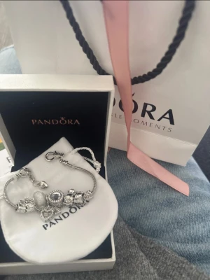 Pandora silverarmband med pärlor - Säljer ett snyggt silverarmband från Pandora med flera detaljerade berlocker och pärlor. Armbandet har klassisk kedja och charmig design med olika former på berlockerna, bland annat hjärta och glittrande stenar. Perfekt accessoar för att lyfta din stil. Vet inte om den är äkta 